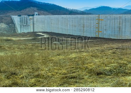 Winter Landscape Of Field Of Dry Grass  In Front Of Concrete Dam With Yellow And Black Water Level R