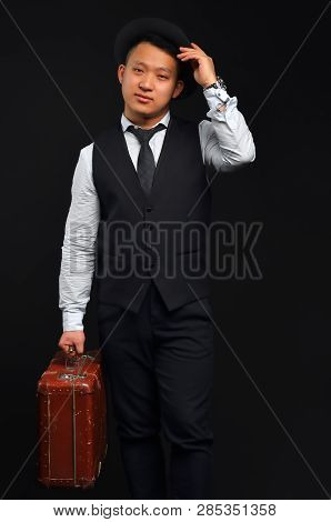 The Photo Of An Asian Man In Full Height, He Carries A Brown Suitcase And Touches His Hat