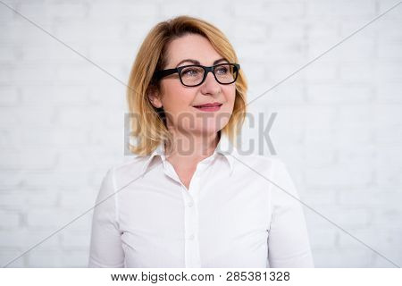 Close Up Portrait Of Thoughtful Cheerful Mature Business Woman In Eyeglasses Posing Over White Brick