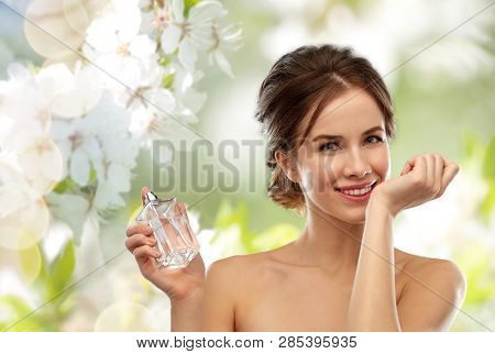 perfumery, beauty and luxury concept - happy smiling young woman smelling perfume from her wrist over natural spring cherry blossom background
