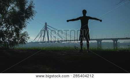 A Silhouette Of A Young Man Performing Exercises Circular Rotations Hands At The Open Air. In The Ba
