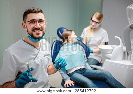 Happy Male Dentist Look On Camera And Smile. He Hold Tools. Female Dentist Stand At Girl. Child Sit 