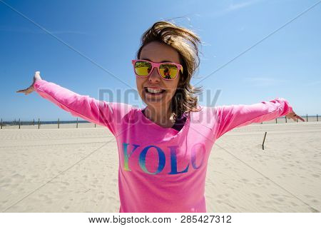 Young Woman Stands On The Beach, Wearing A Neon Pink Yolo Shirt With Sunglasses, As Her Hair Blows I