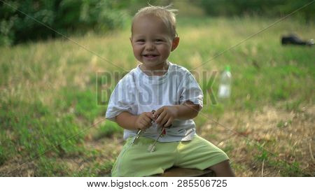 Baby With Smile Sitting In The Park With Green Grass And Trees. Happy Boy Child Playing With Keys. B