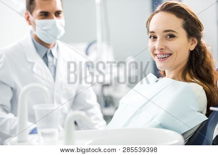 Selective Focus Of Woman In Braces Smiling With Dentist Standing In Mask On Background