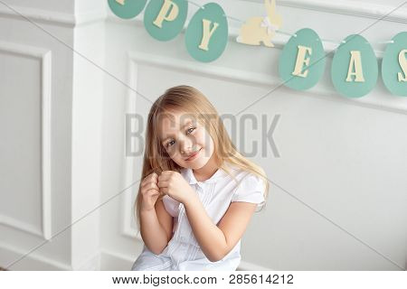 Cute Little Blonde Girl Sitting In A Bright Room Near The Window On The Background Of Streamers With