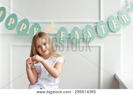 Cute Little Blonde Girl Sitting In A Bright Room Near The Window On The Background Of Streamers With