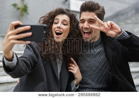 Image of satisfied couple man and woman 20s in warm clothes taking selfie photo on cell phone while sitting on stairs outdoor