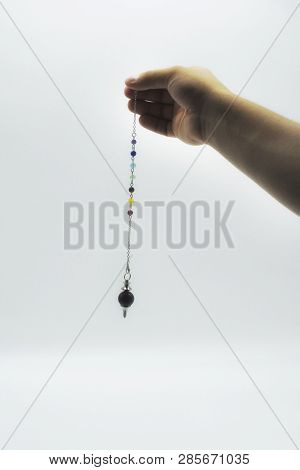 Pendulum Dowsing On An Isolated White Background With An Amethyst Crystal And Colored Chakra Stones.