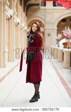 Full body portrait. Young beautiful woman in stylish in a long burgundy coat, indoor shop