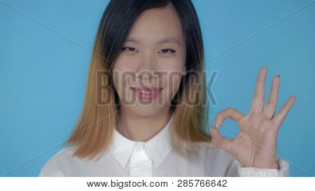 Close Up Portrait Young Asian Woman Laughing Shows Sign Symbol Ok On Blue Background In Studio. Attr