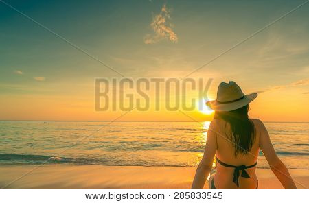 Back View Of Asian Woman Wear Bikini And Straw Hat Sit On Sand Beach Relax And Enjoy Holiday At Trop
