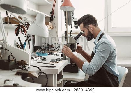 Very Close. Side View Of A Male Jeweler Looking At The New Jewelery Product Through Microscope In A 