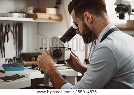 Focused On A Process. Male Jeweler Looking At The Ring Through Microscope In A Workshop.
