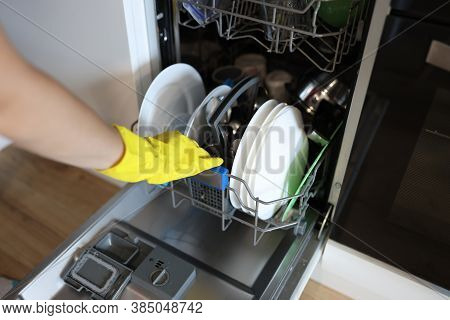 Womans Hand In Rubber Glove Puts Dishes Into Dishwasher. Care, Operation And Maintenance Concept.