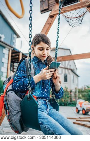 Girl Listening To Music On Her Phone While Sitting On A Swing With A Backpack Behind Her Back.