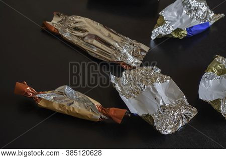 Random Blank Candy Wrappers On A Black Table. Multicolored Foil Wrappers With Crumbs And Spots. Choc