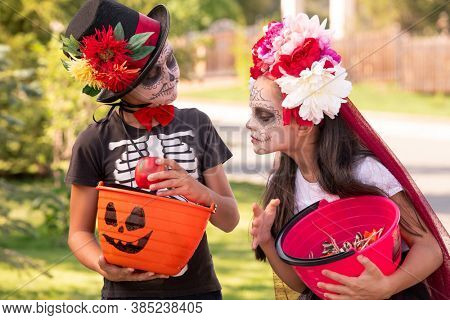 Cute halloweeen girl with dark long hair looking at basket with treats in hands of her friend holding red ripe apple while boasting with it