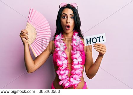 Beautiful hispanic woman wearing bikini and handfan holding hot word afraid and shocked with surprise and amazed expression, fear and excited face. 