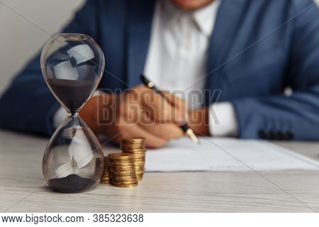 Businessman Approves Important Contract In Office. Stack Of Coins And Hourglass On Desk Close-up. Ti