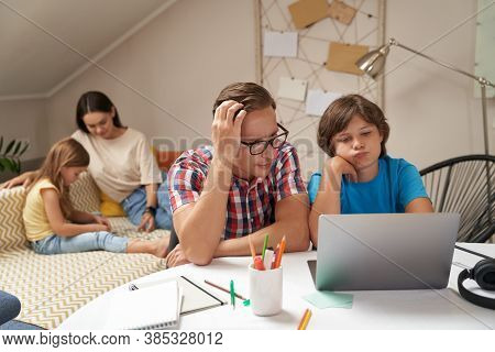 Worried Father Helping His Little Son With Homework While Mother And Daughter Sitting On Sofa And Pl