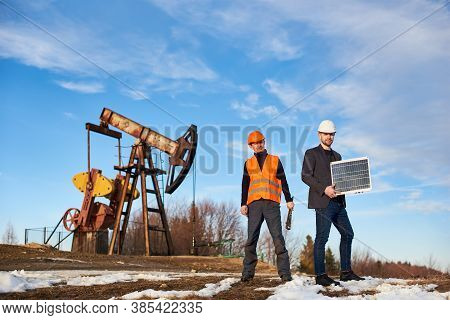 Two Men On Oil Field With Pump Jack On Background. Businessman Holding Portable Solar Panel, Oil Wel