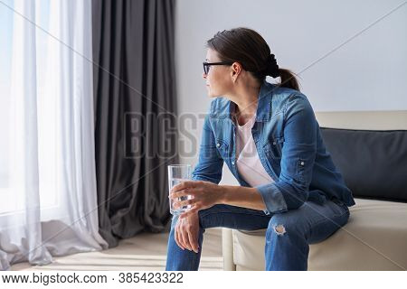 Serious Sad Middle Aged Mature Woman Sitting At Home Holding Glass Of Water Looking Out The Window. 