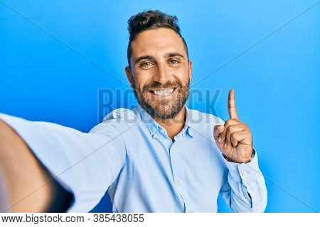Handsome man with beard taking a selfie photo with smartphone surprised with an idea or question pointing finger with happy face, number one 