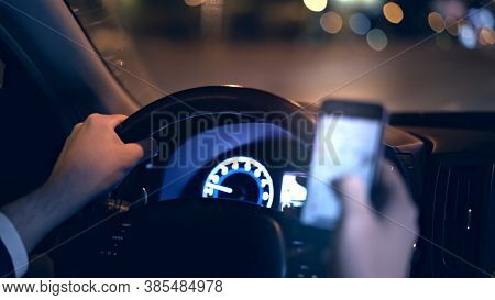 Male Hands On Steering Wheel, Backseat Shot With View On Road At Night. Hands On Wheel With Smartpho