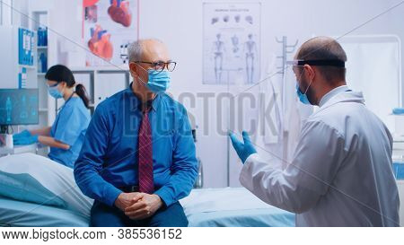 Senior Man At Doctor Appointment During Covid-19 Pandemic. Patient Wearing Mask And Doctor In Protec