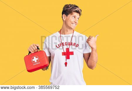 Young handsome man wearing lifeguard t shirt holding first aid kit pointing thumb up to the side smiling happy with open mouth 
