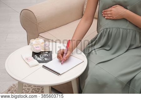 Pregnant Woman With Sonogram And Baby's Accessories Writing Names List At Small White Table, Closeup