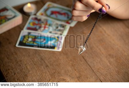 Arm Of A Dowser With Hand-held Pendulum On The Background Of A Burning Candle And Tarot Cards. Selec