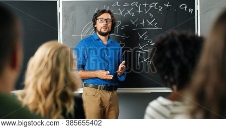 Caucasian Man Teacher At School At Blackboard Talking With Pupils Or Students And Asking Question. M