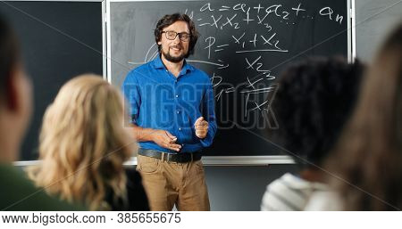 Caucasian Man Teacher In Glasses At School At Blackboard Talking With Pupils Or Students. Math Class