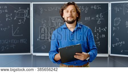 Portrait Of Caucasian Male Teacher Standing In Classroom With Folder In Hands Looking At Camera. Mat