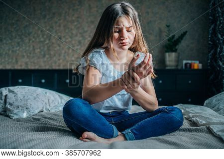 A Young Brunette Girl Is Sitting On A Bed In Her Room. She Holds On To A Sore Hand. Horizontal Shot,