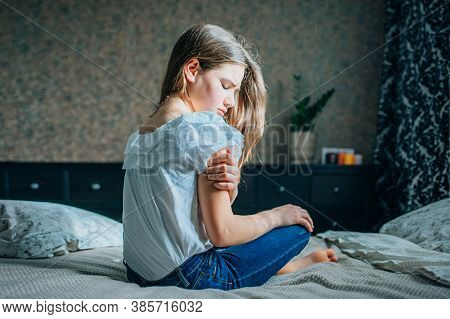 Young Brunette Girl Sitting On The Bed In Her Room, Holds An Injured Shoulder With Her Hand, Horizon