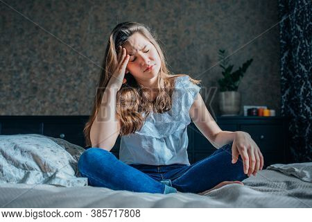 A Sad Dark-haired Girl In A White Blouse And Blue Jeans Is Sitting On The Bed In Her Bedroom.