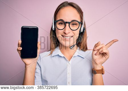Beautiful call center agent woman working using headset holding smartphone holding screen very happy pointing with hand and finger to the side