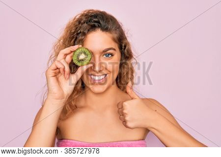 Beautiful woman with blue eyes wearing towel shower after bath holding kiwi fruit over eye happy with big smile doing ok sign, thumb up with fingers, excellent sign
