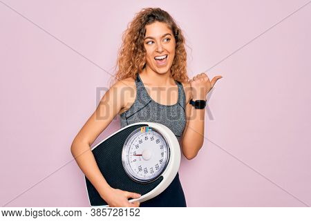 Young beautiful slim sporty woman with blue eyes controlling weight holding weighting machine pointing and showing with thumb up to the side with happy face smiling