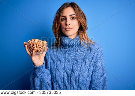 Young beautiful brunette woman holding bowl with peanuts over blue background with a confident expression on smart face thinking serious