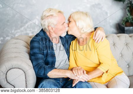 Elderly Gray-haired Couple In Love Is Kissing. Senior Spouses Sit On Cozy Sofa At Home In Embrases