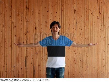 Man Wearing Estonia Flag Color Shirt And Standing With Arms Wide Open On The Wooden Wall Background,