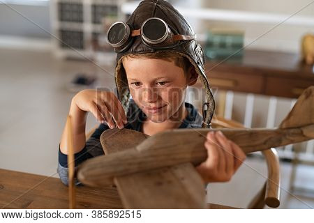 Boy wearing pilot helmet playing with vintage wooden airplane at home. Happy child playing with wooden flight simulator. Little boy holding handmade plane with pilot cap and goggles at home.