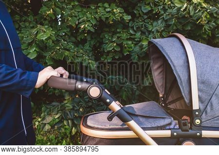 Mom In Blue Coat Walking With Her Baby In Park With Grey Stroller. Nature Background. Side View. Clo