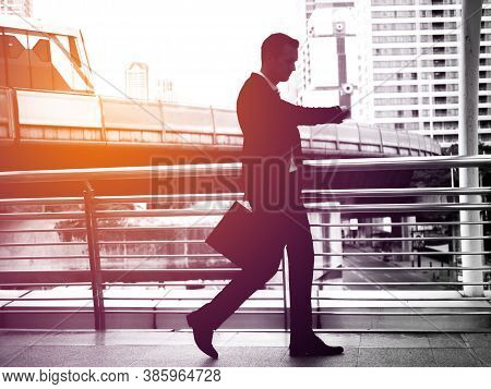Businessman Walking On The Skywalk Hastily. In His Hand Is A Black Leather Briefcase. With Flare Lig