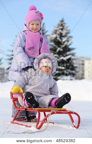Bebê vestido com roupas quentes, senta-se em vermelhos trenó e irmã mais nova de stands atrás no inverno.