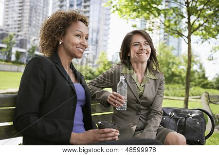 Due imprenditrici sorridente seduto sulla panchina del parco con tazza di caffè e borraccia